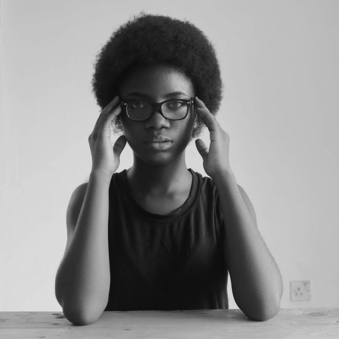 Black and white portrait of a young woman wearing glasses, seated indoors.